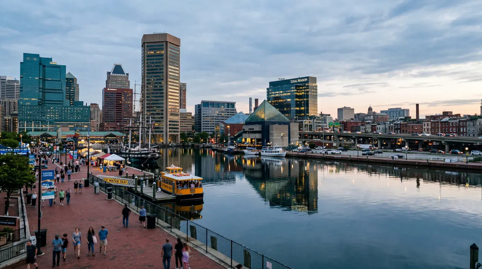 Baltimore Maryland Inner Harbor waterfront, modern buildings reflected in water, the edge of the tourist zone visible, urban reality just beyond. documentary travel photography, realistic, no text, no watermark, 16:9
