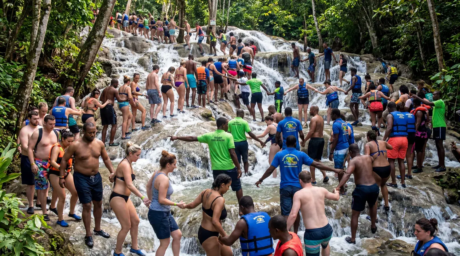 Dunn's River Falls Jamaica overcrowded with tourists climbing in a human chain, guides directing hundreds of people up the waterfall, crowded scene, documentary travel photography, photorealistic, no text, no watermark, 16:9