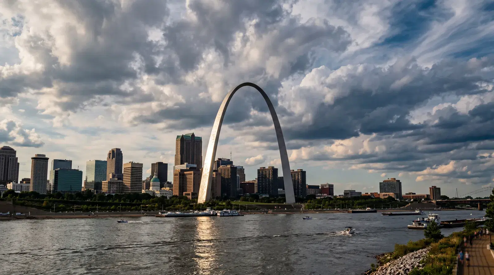 St Louis Missouri Gateway Arch gleaming in afternoon sun, Mississippi River in foreground, dramatic clouds overhead, city skyline behind. documentary travel photography, realistic, no text, no watermark, 16:9