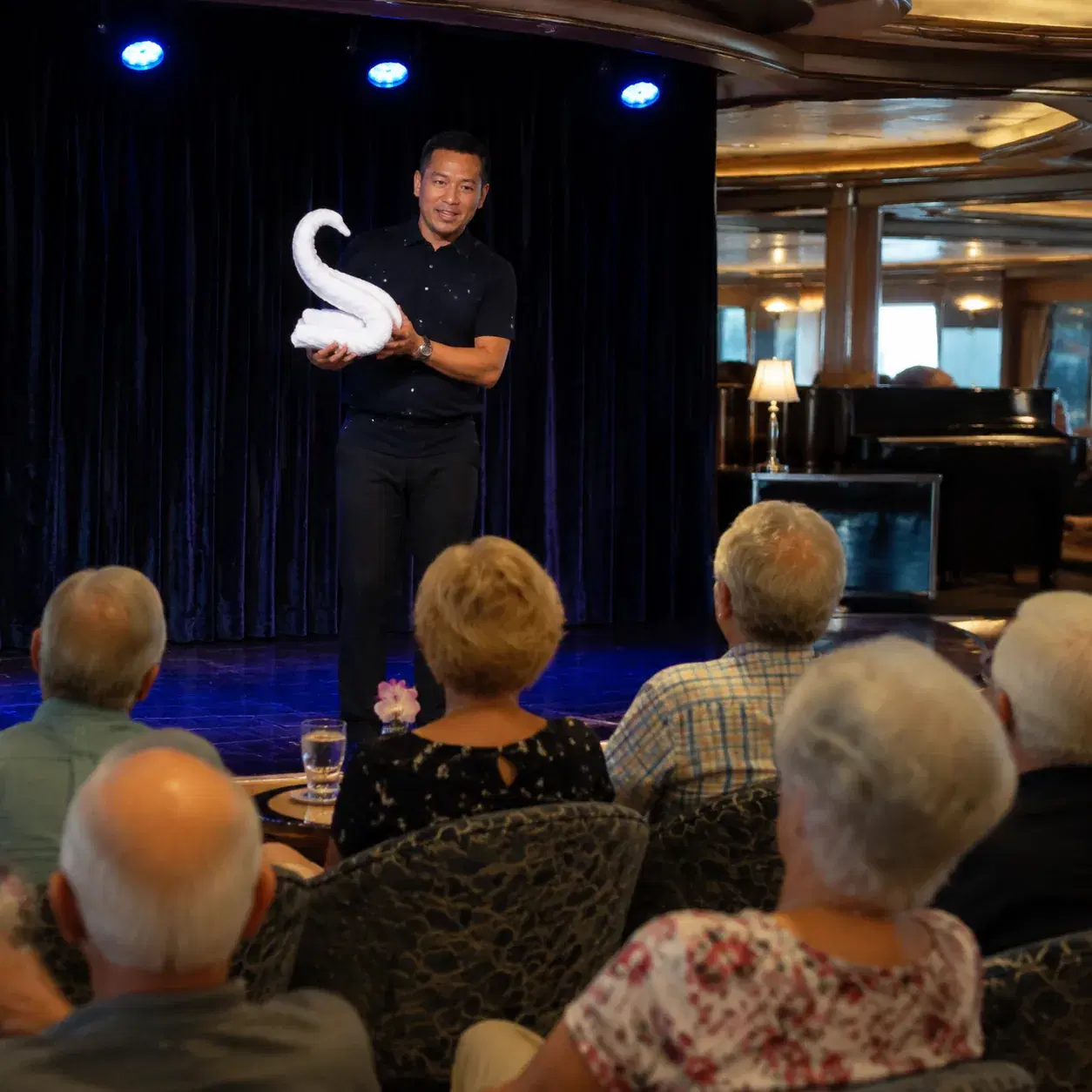 Realistic editorial photo of cruise lounge karaoke setup with microphone, small stage, seated passengers, and warm color