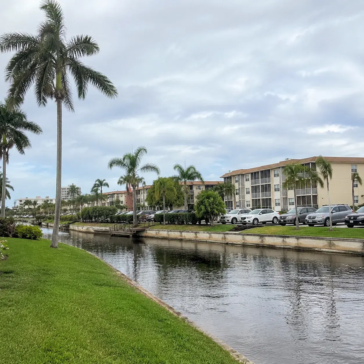 Realistic editorial photo of an older Deerfield Beach retirement condo area with canals, parked cars, palm trees, and so