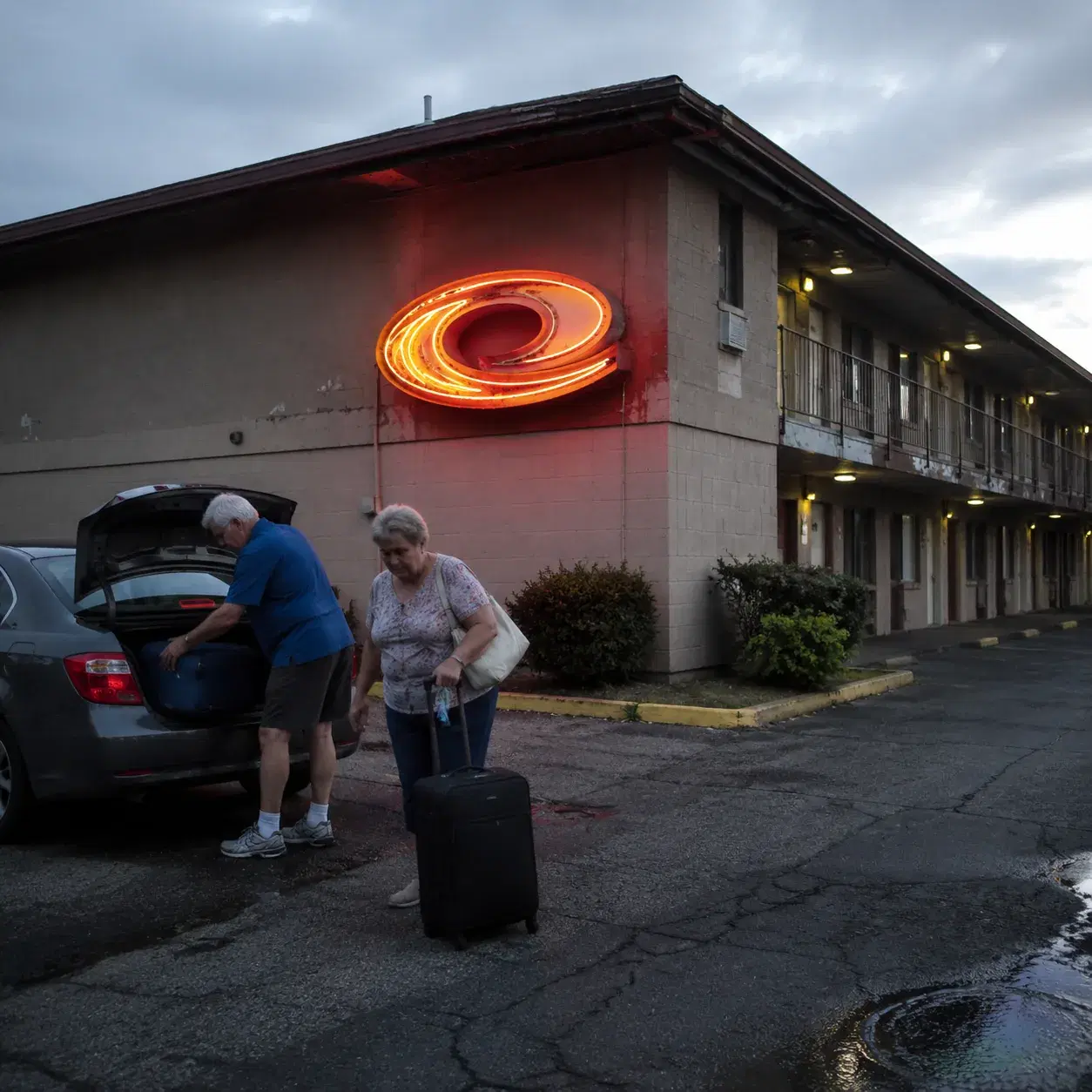 Worn Econo Lodge exterior with flickering neon sign, cracked parking lot, two older American tourists loading luggage wi