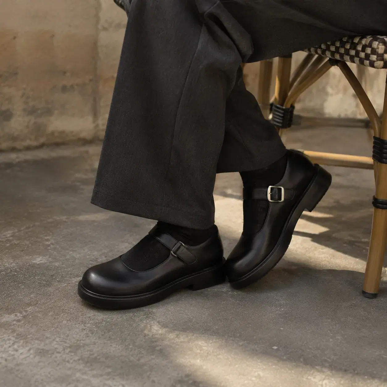 Realistic editorial photo of heavy rounded-toe Mary Jane shoes worn with trousers beside a cafe chair, thick strap and s
