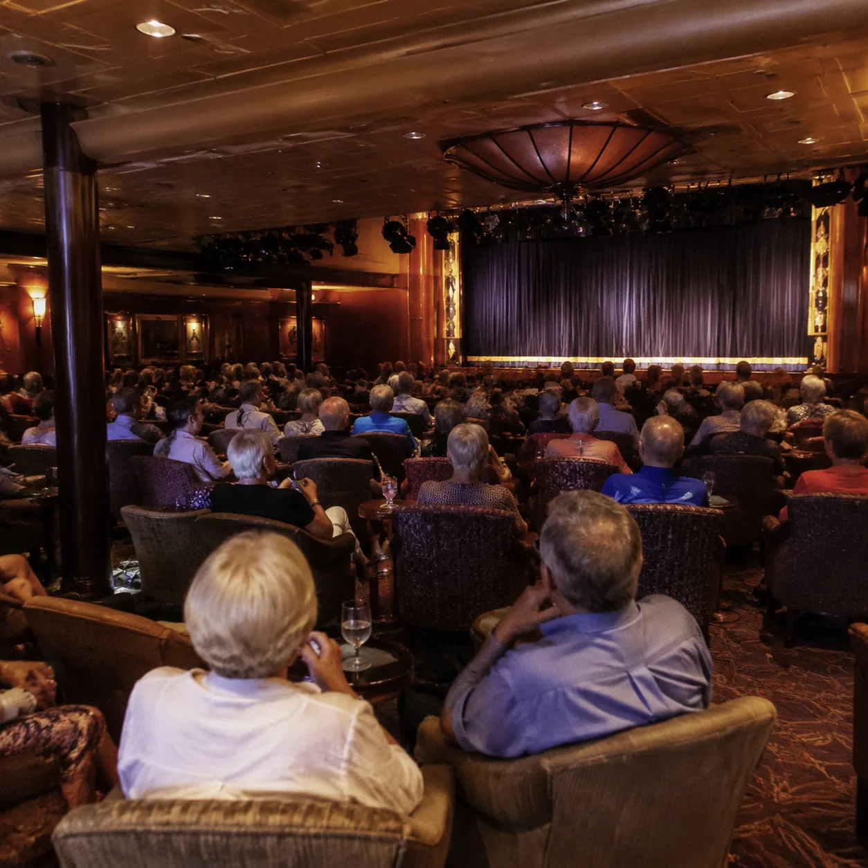 Older British-style cruise ship lounge with mature passengers seated in rows before an evening show, classic decor