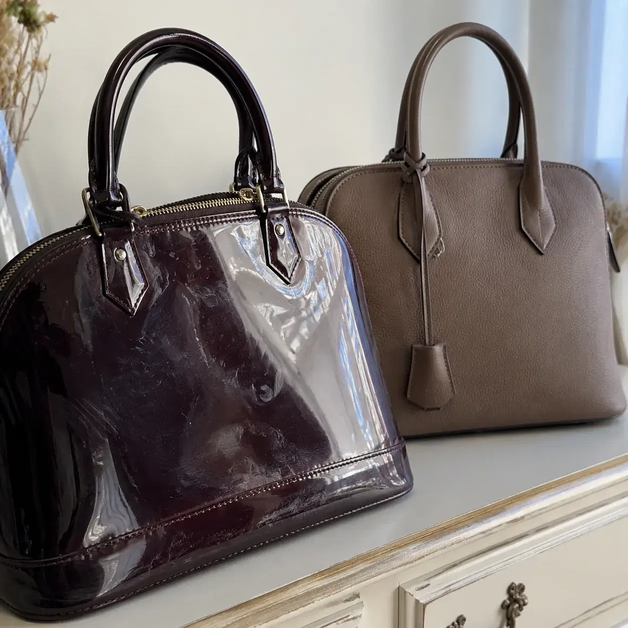 Realistic editorial close-up photo of a glossy patent handbag with visible scuffs and fingerprints beside a matte leathe