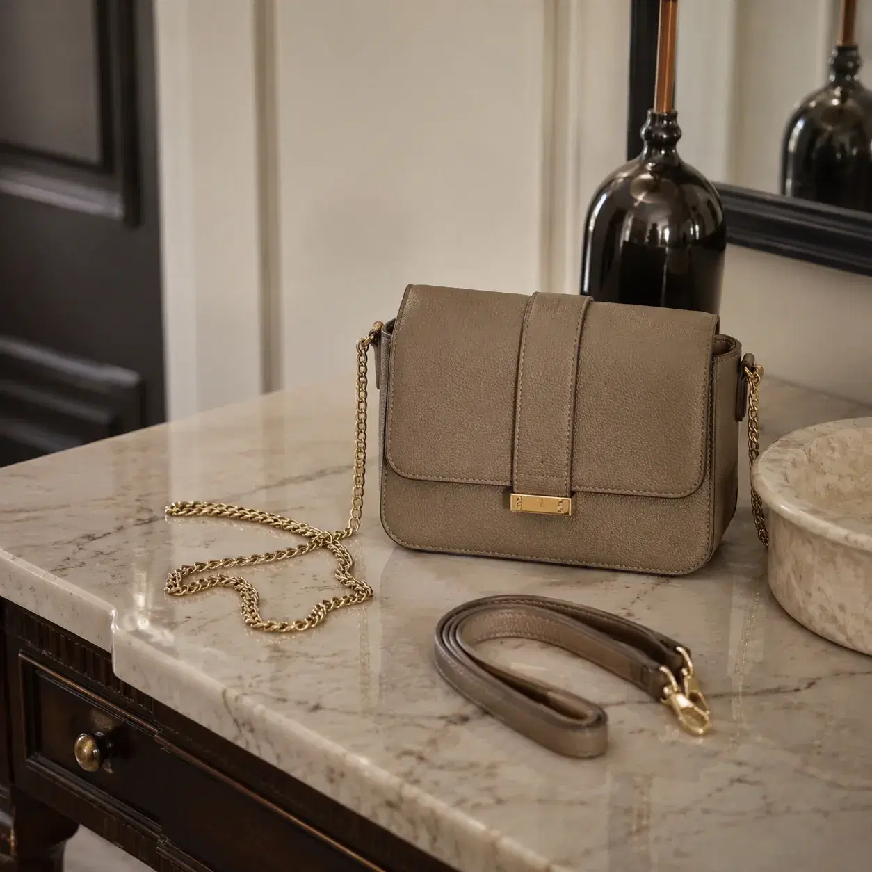 Realistic editorial photo of a handbag with a thin cheap chain strap kinked on a marble entry table beside a smoother le
