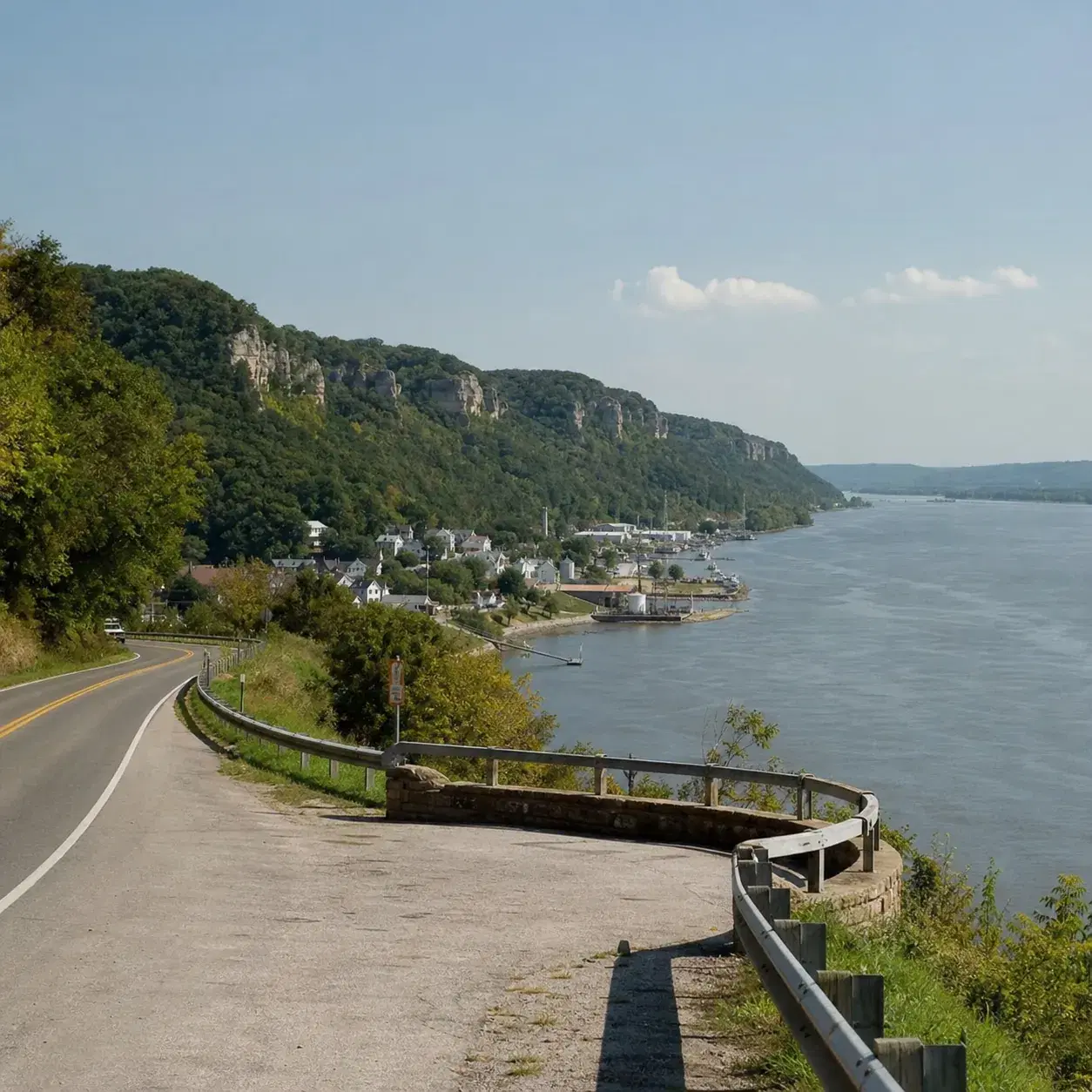 Realistic editorial photo of a river road beside the Mississippi with bluffs, a small riverside town, and a classic road