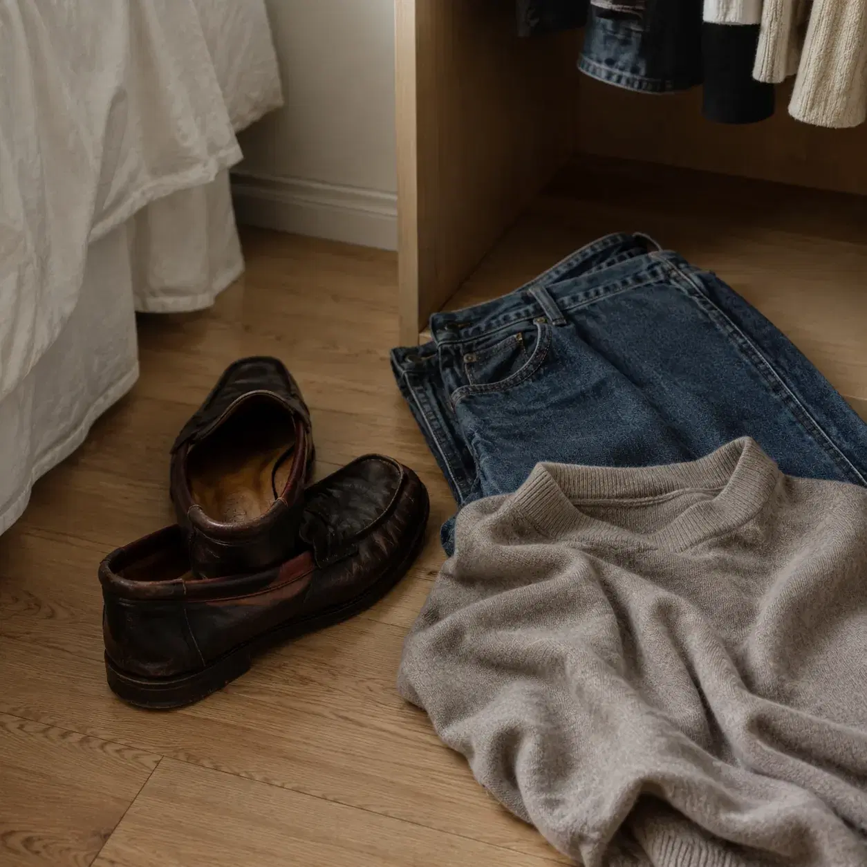Realistic editorial photo of collapsed leather loafers beside neat straight-leg jeans and a sweater on a closet floor, w