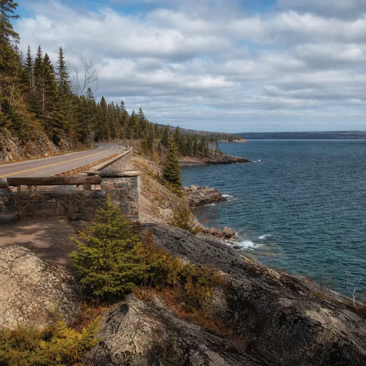 Realistic editorial photo of a Minnesota lakefront highway near Lake Superior with rocky shoreline, evergreen trees, and