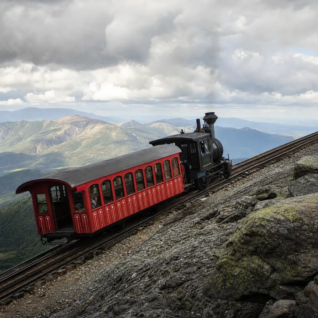 Realistic editorial photo of the Mount Washington Cog Railway climbing a steep rocky mountain slope in New Hampshire wit