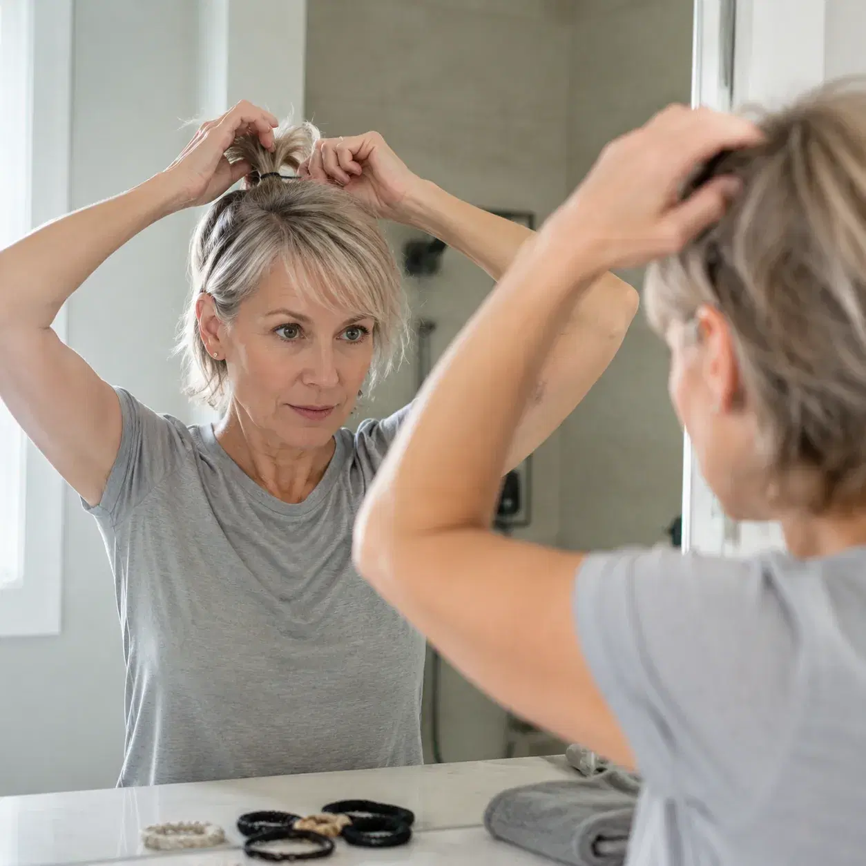 Realistic editorial photo of a woman over 50 trying to pull freshly washed short layers into a small ponytail, bathroom