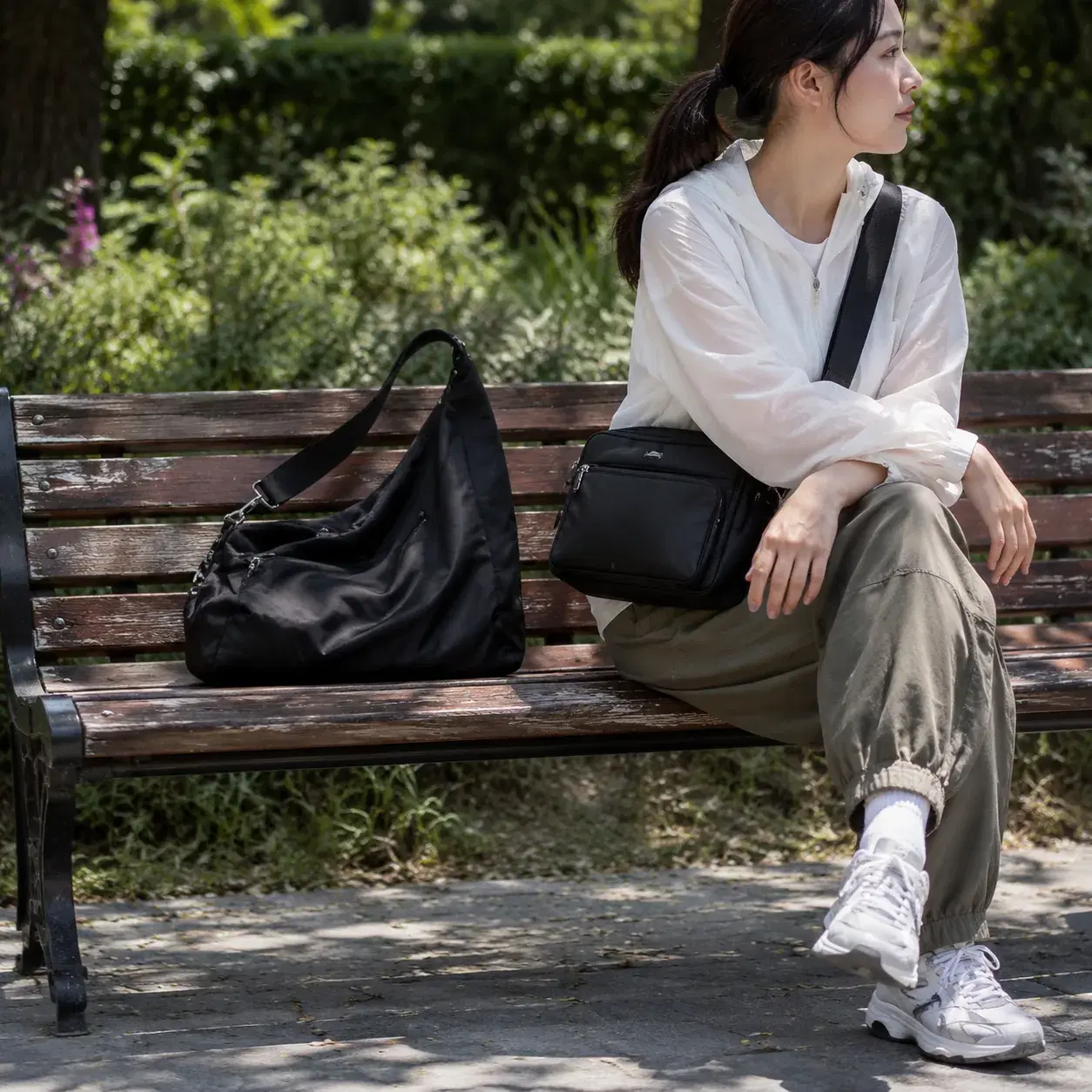 Realistic editorial photo of a casual nylon shoulder bag slumping on a park bench beside a cleaner structured nylon cros