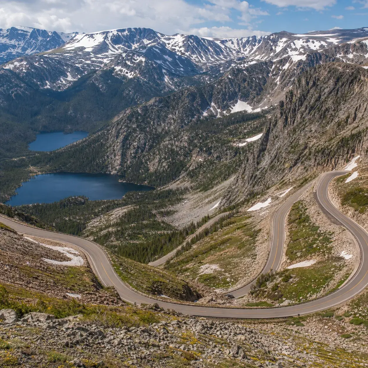 Realistic editorial photo of a high alpine highway near Beartooth Pass with switchbacks, mountain lakes, and patches of 