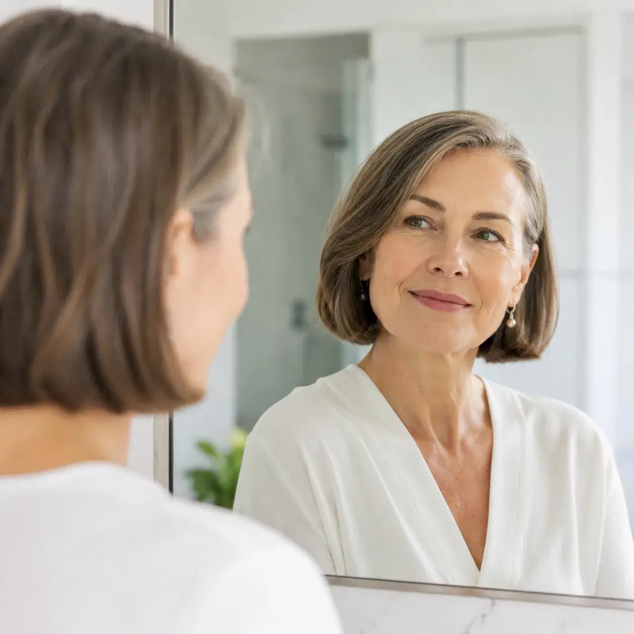 Realistic editorial photo of a woman over 50 with a bob haircut tucked behind one ear, simple earrings, clean bathroom m