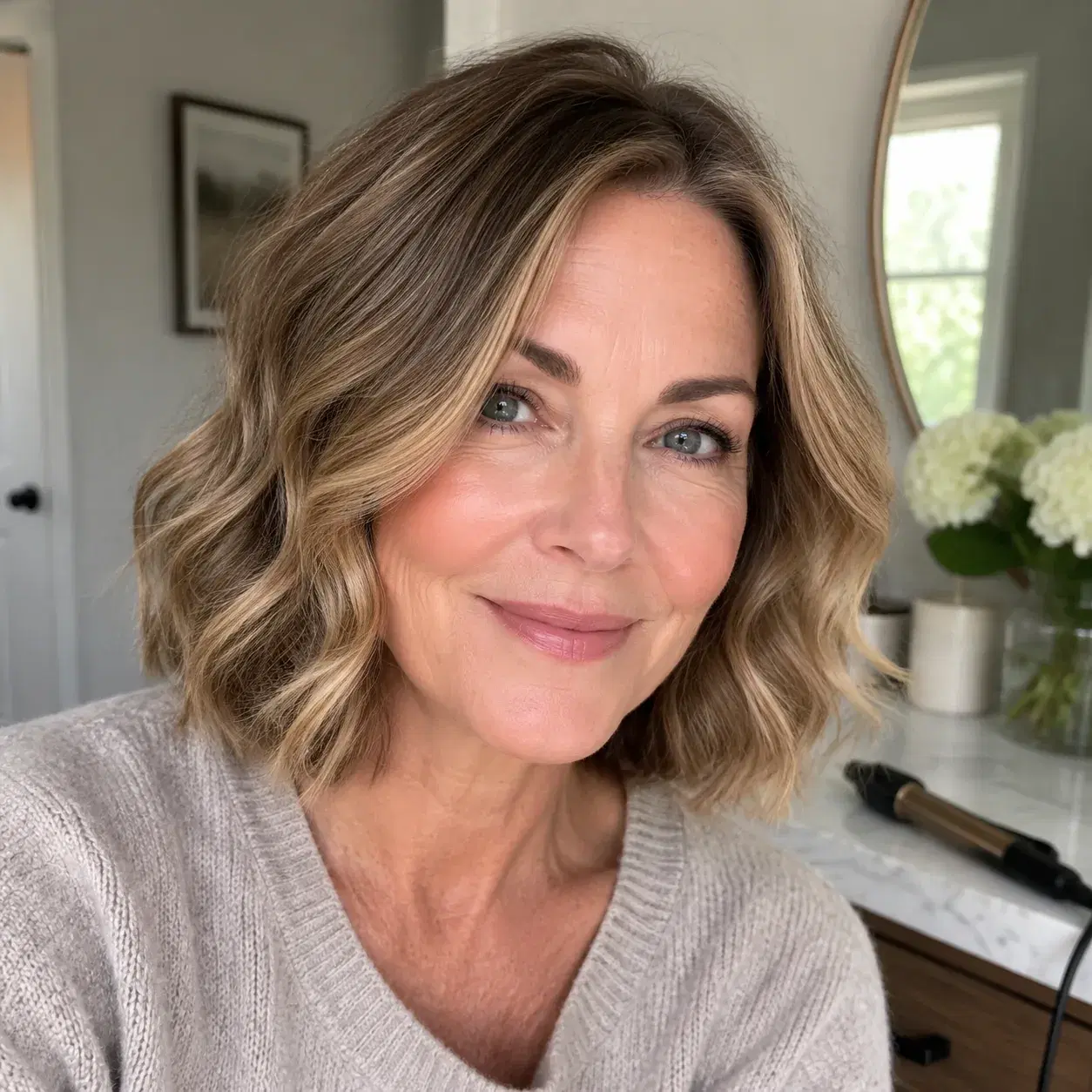 Realistic editorial photo of a woman over 50 with a wavy bob and blunt bottom line, casual home vanity setting with curl