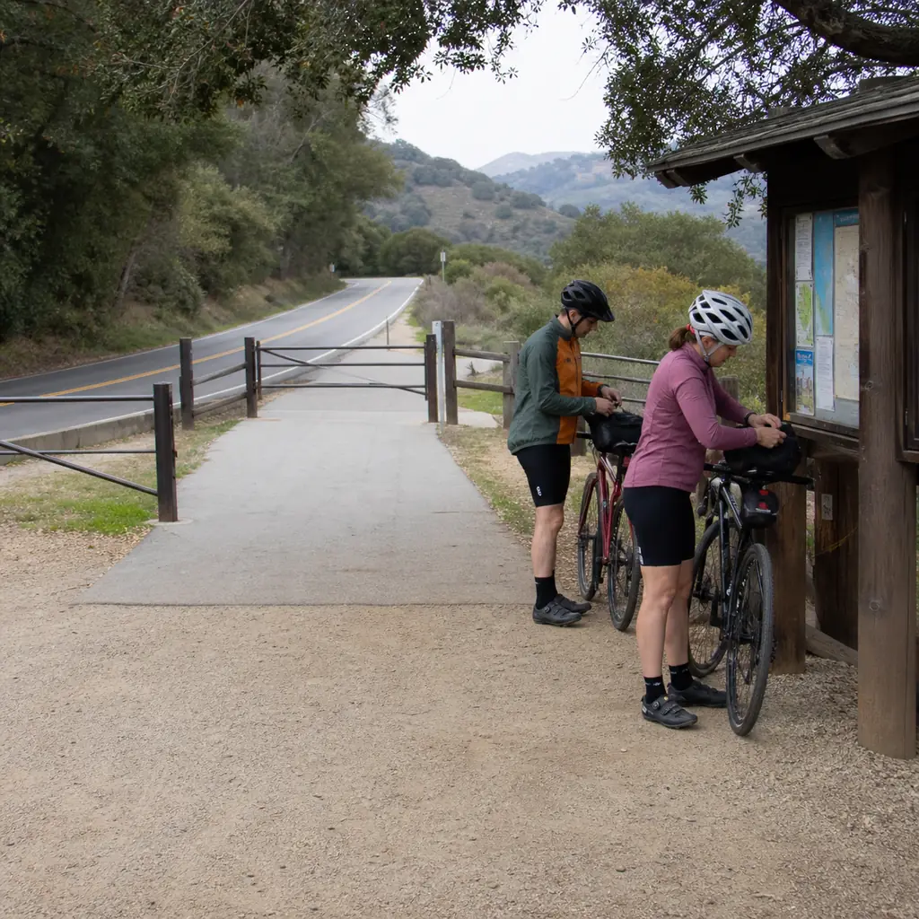 Realistic editorial photo of two cyclists arranging bikes and helmets neatly beside a trail kiosk while keeping the gate