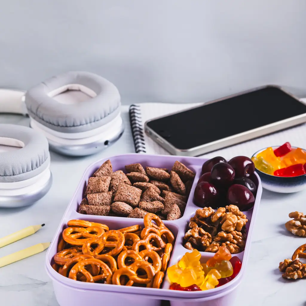 A divided snack box with nuts, pretzels and fruit beside travel essentials