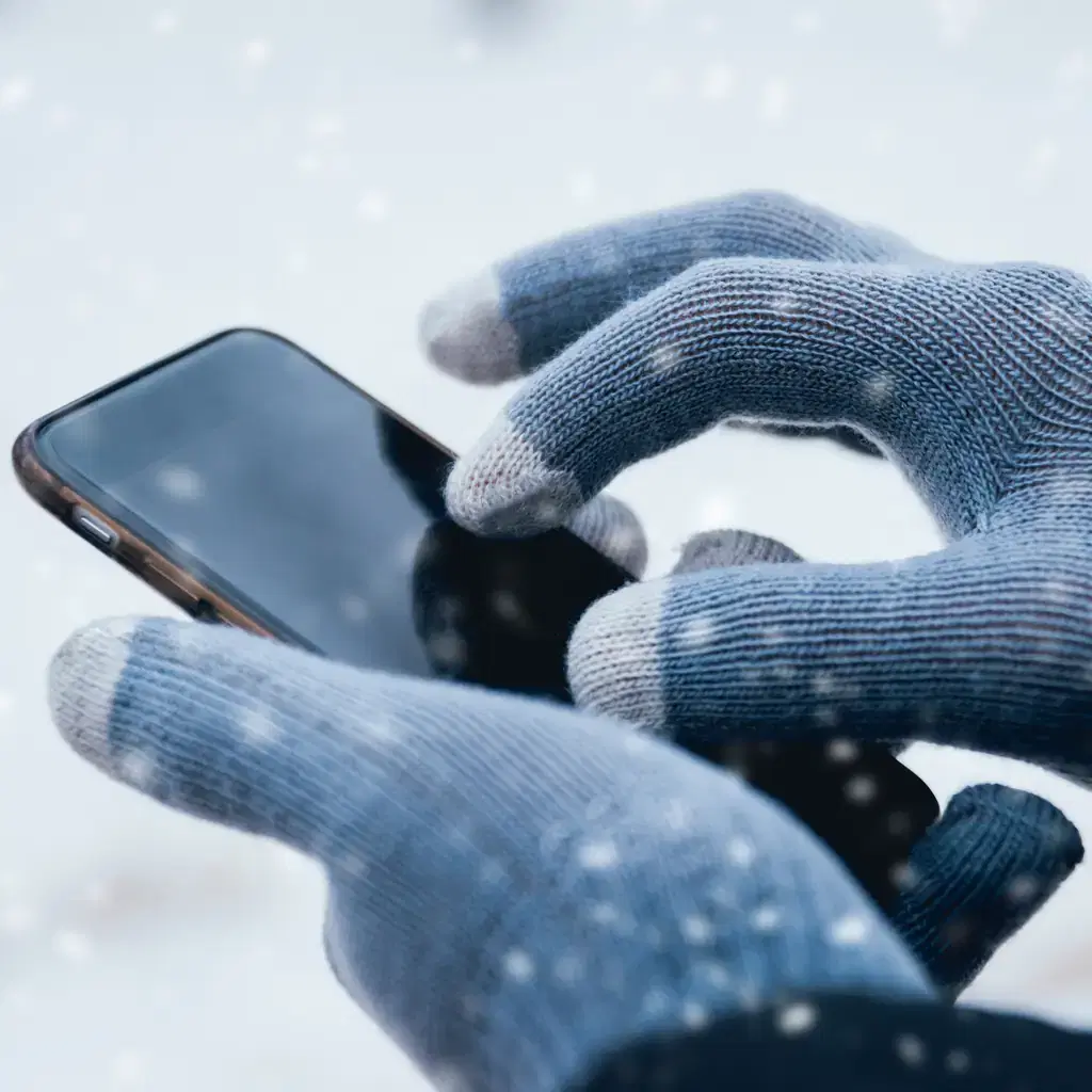 Hands in thin knit liner gloves using a smartphone outdoors in the snow, thick gloves held in the other hand