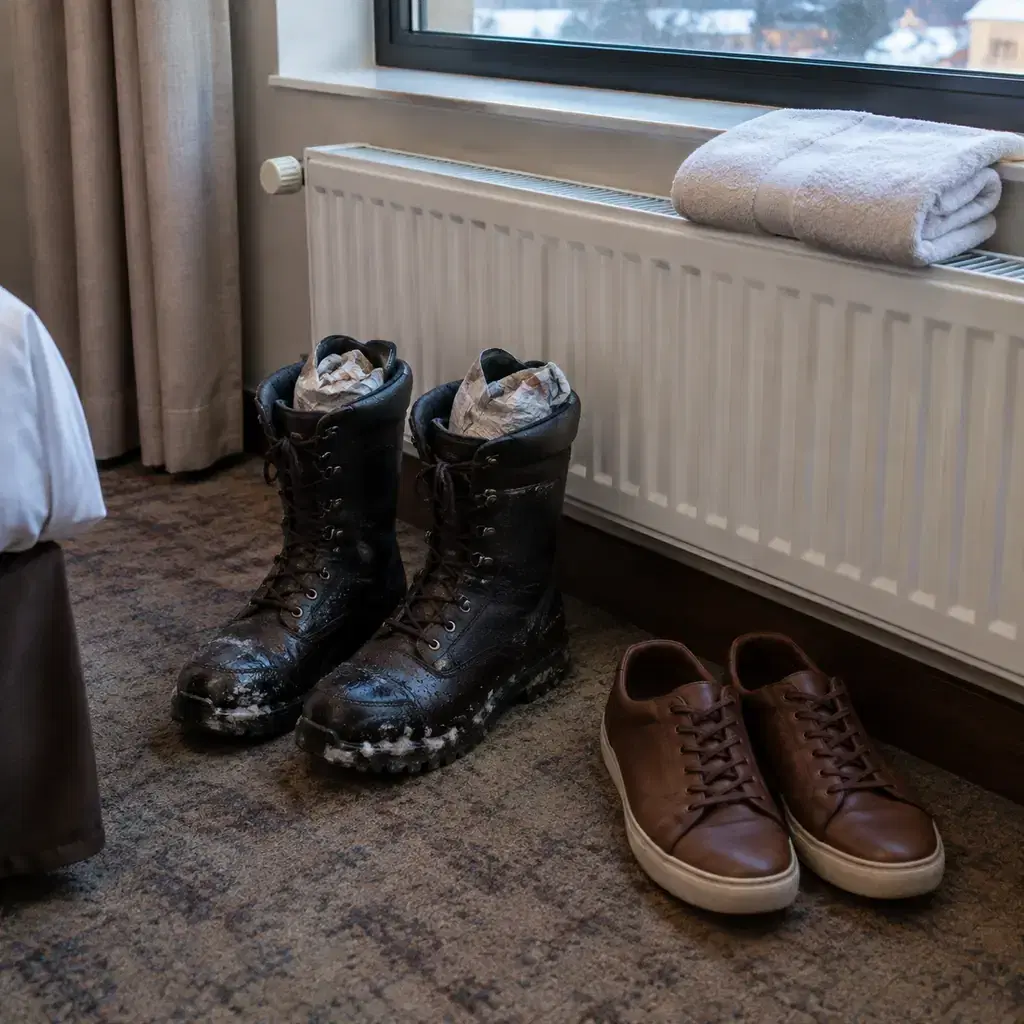 Wet winter boots stuffed with newspaper drying near a hotel radiator, with a dry second pair of shoes beside them