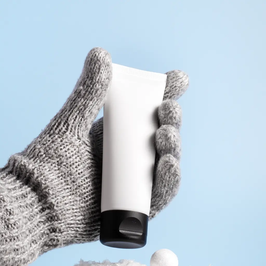 A knit-gloved hand holding a small tube of hand cream against snow and blue sky