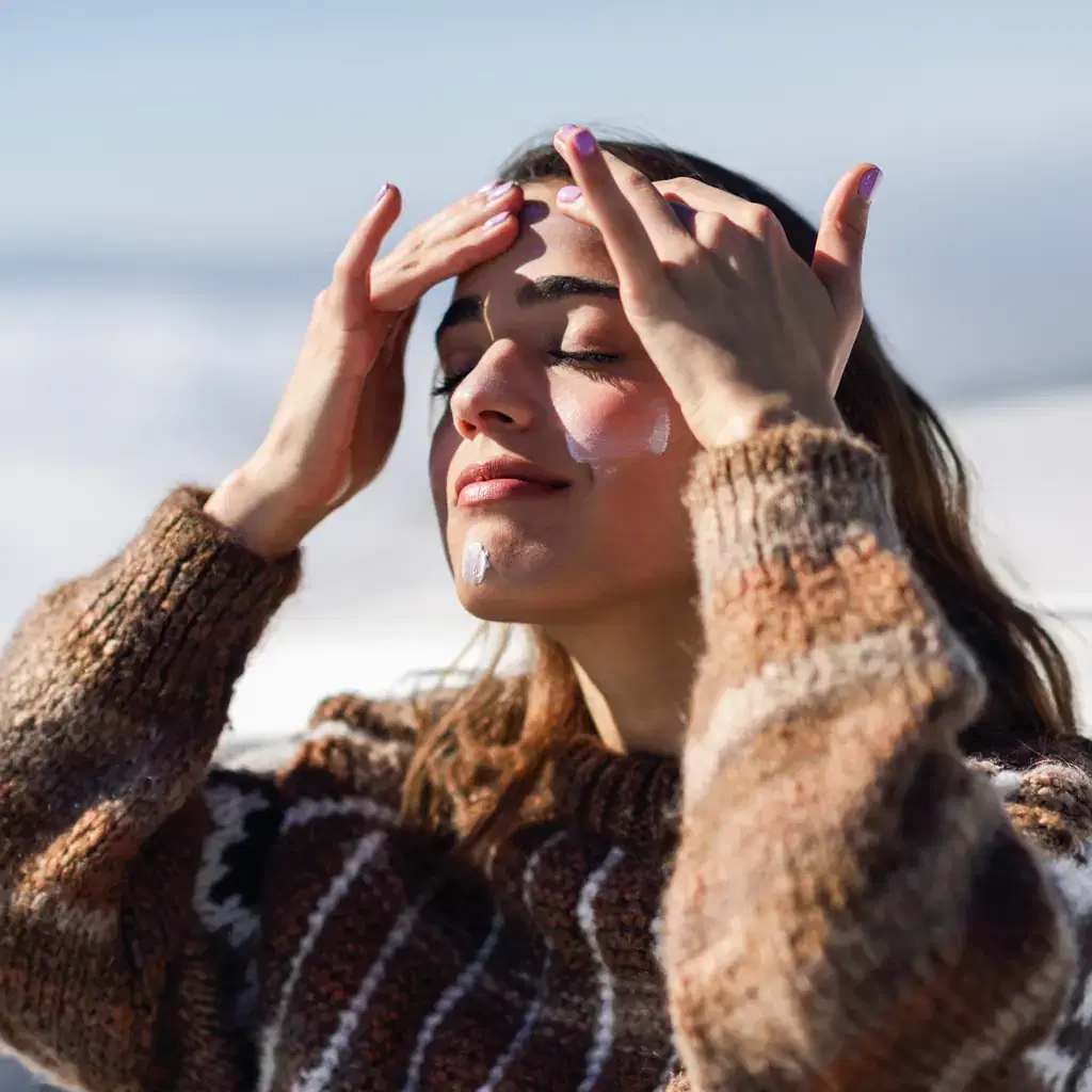 A person in winter clothing applying sunscreen to their face on a bright snowy slope while holding sunglasses
