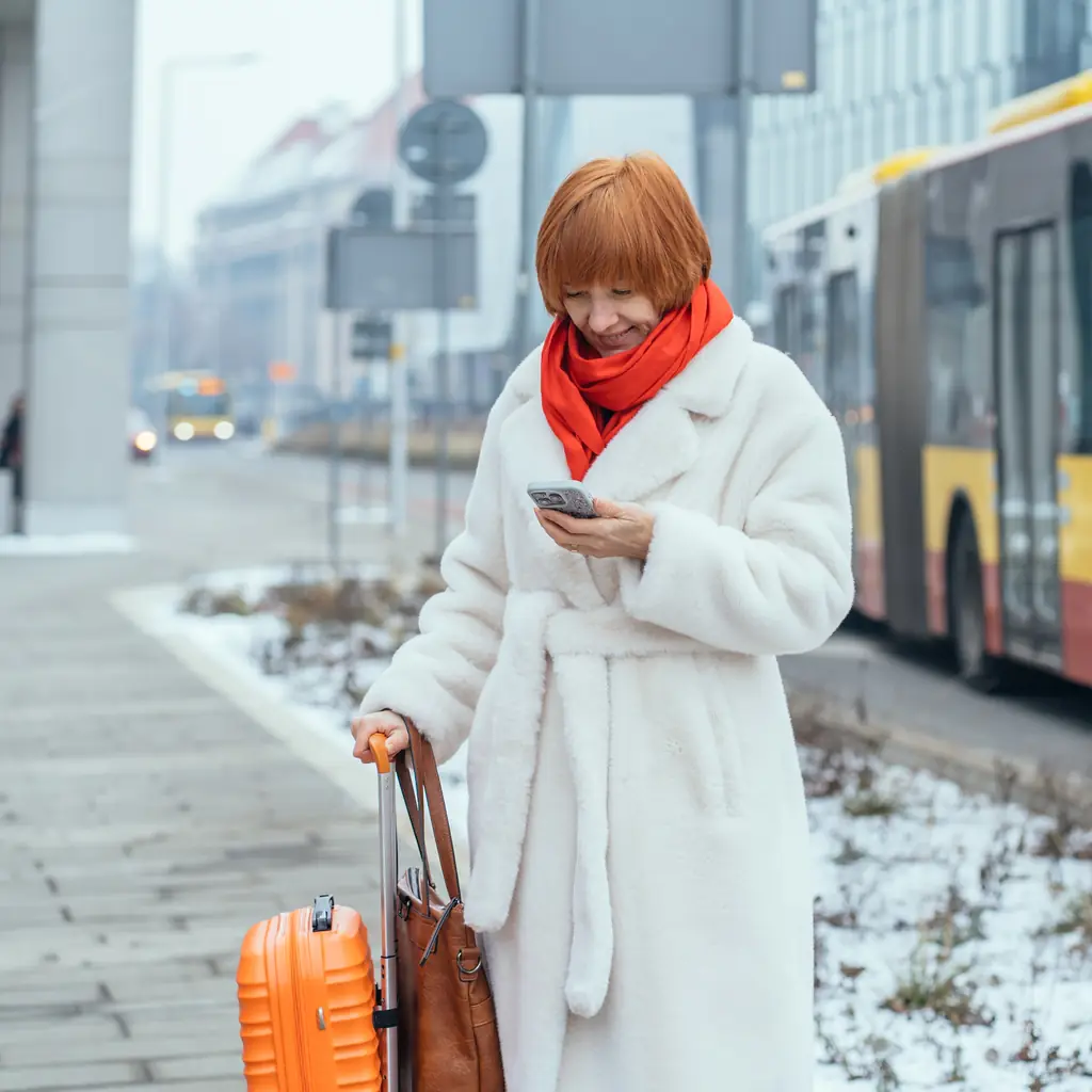 A traveler in a winter coat checking a phone near an airport departures board while snow falls outside