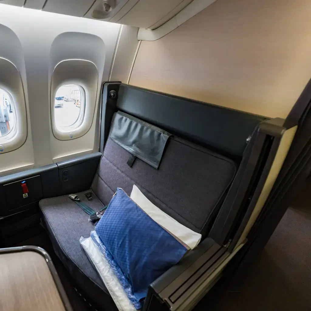 A blue pillow and folded white bedding laid on a business class suite seat beside the cabin windows
