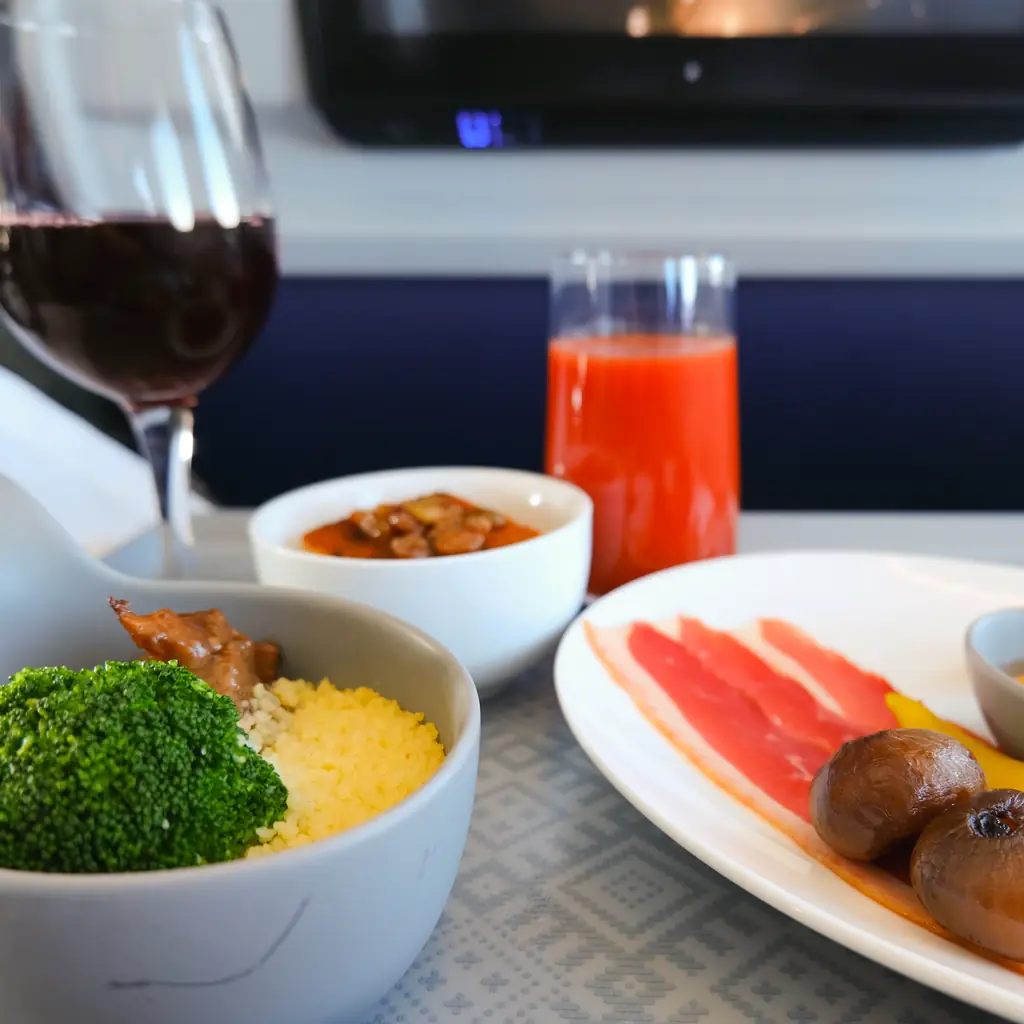 A business class meal of plated starters and a main with a glass of red wine and tomato juice on a tray table
