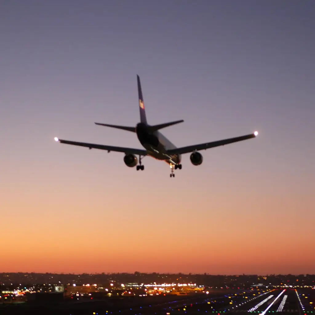 An airliner with its lights on flying low over runway lights against an orange dusk sky