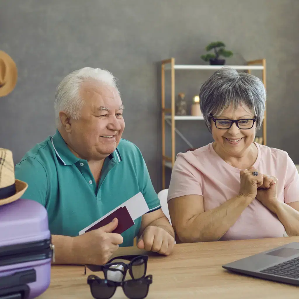 A smiling older couple at a table with a laptop, passport and packed suitcase while planning a trip