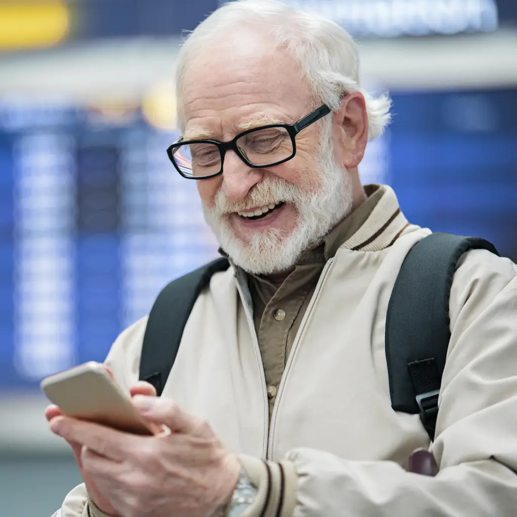 An older traveler with a backpack smiling at his smartphone in an airport terminal, the departures board blurred behind
