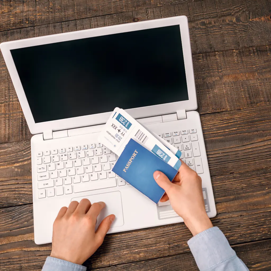 An older adult's hands on a laptop keyboard beside a passport and coffee mug while booking a flight
