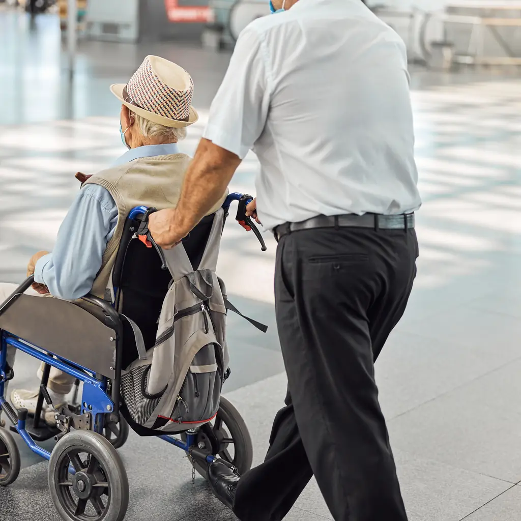 An airport assistance worker pushes an older traveler in a wheelchair along a terminal concourse