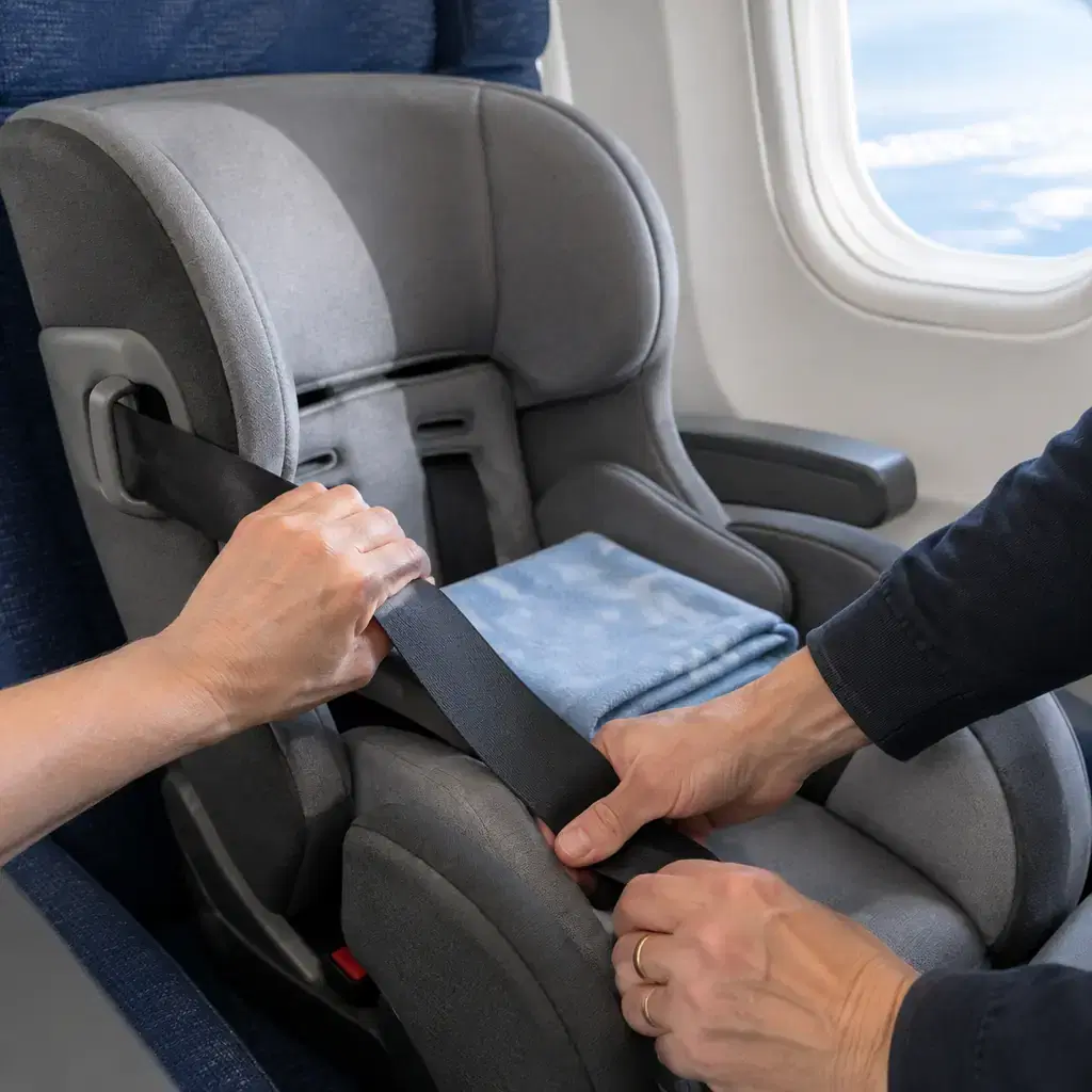 Hands tighten an airplane seatbelt through the base of a child car seat installed in a window seat