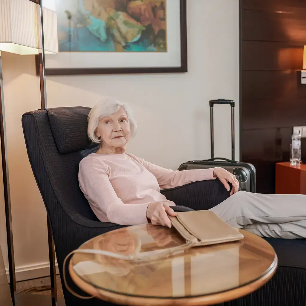 An older traveler rests in an armchair in a quiet guest bedroom beside an open suitcase