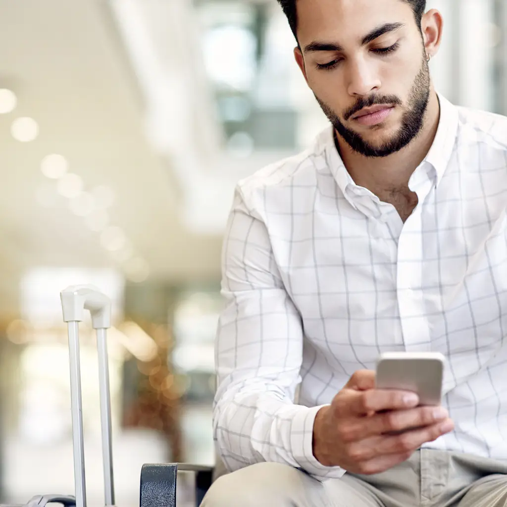 A traveler checking his phone while seated at an airport gate with luggage, illustrating a pre-purchased standalone seat upgrade.