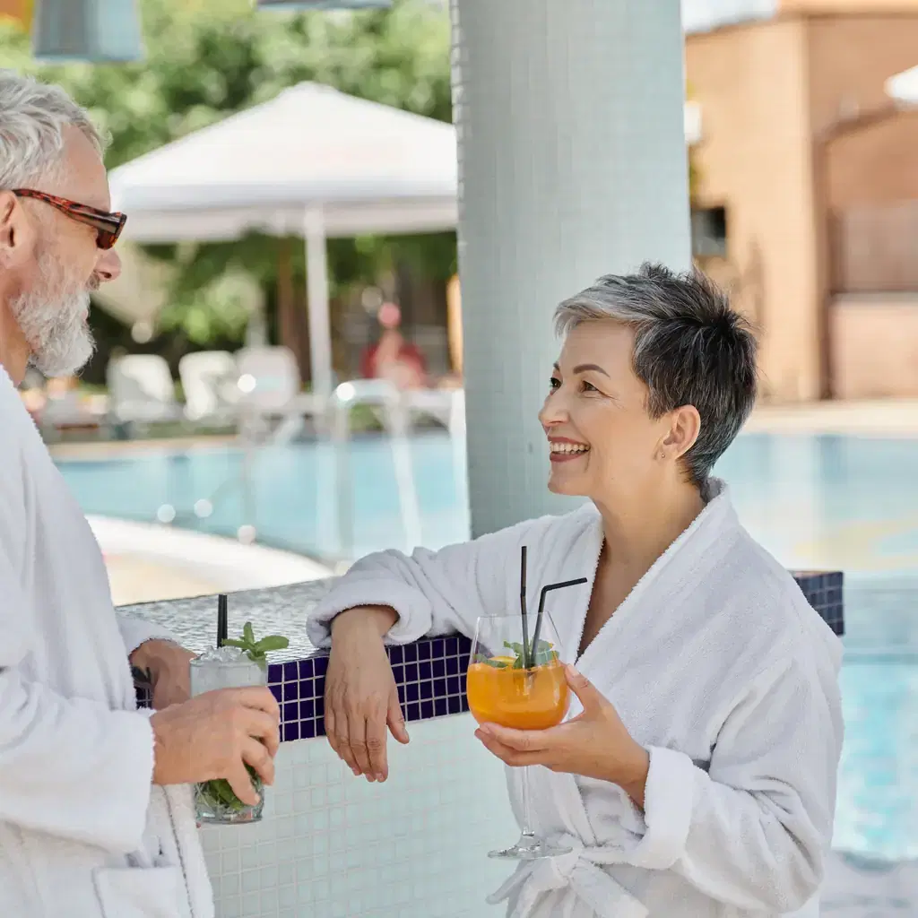 A middle-aged couple in robes enjoying cocktails poolside, illustrating who a fully adults-only property suits.